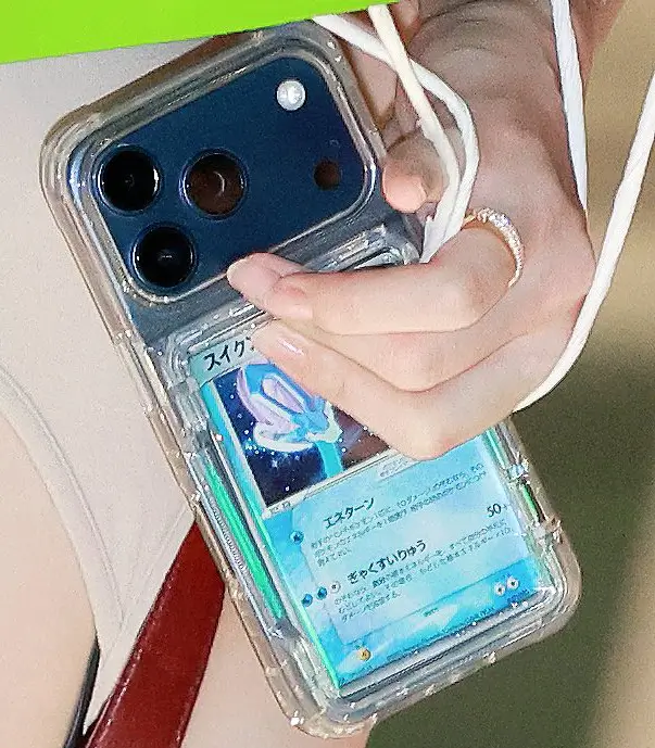 Close-up of a smartphone in a transparent protective case displaying a Japanese Suicune Pokémon card tucked behind the device, held by a hand wearing a silver ring with a white wrist strap attached. The image highlights the growing trend of showcasing collectible trading cards as everyday phone accessories.