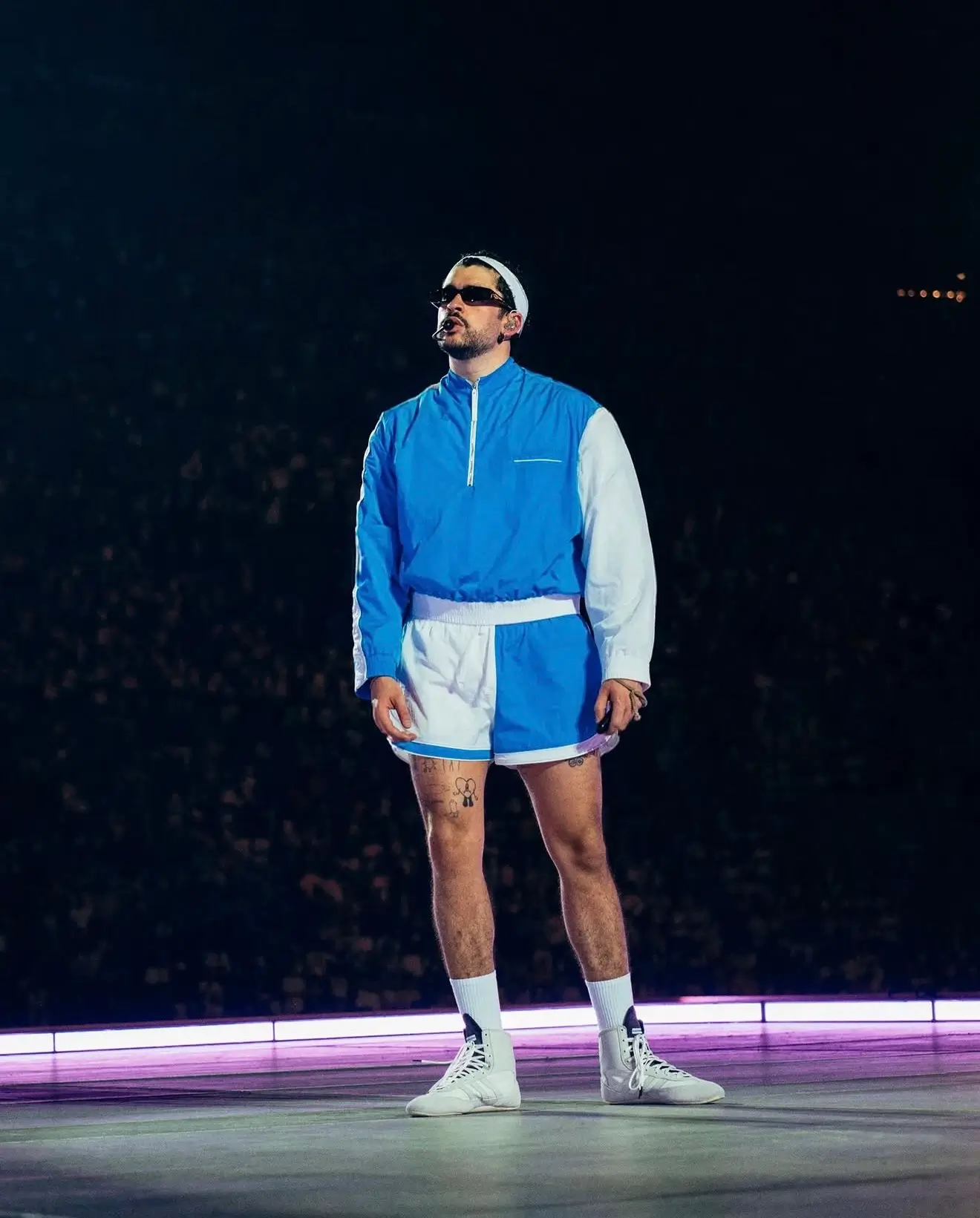 Performer onstage wearing a blue-and-white color-block athletic set featuring a quarter-zip windbreaker, matching running shorts, white headband, crew socks, and white sneakers while performing under arena lights.