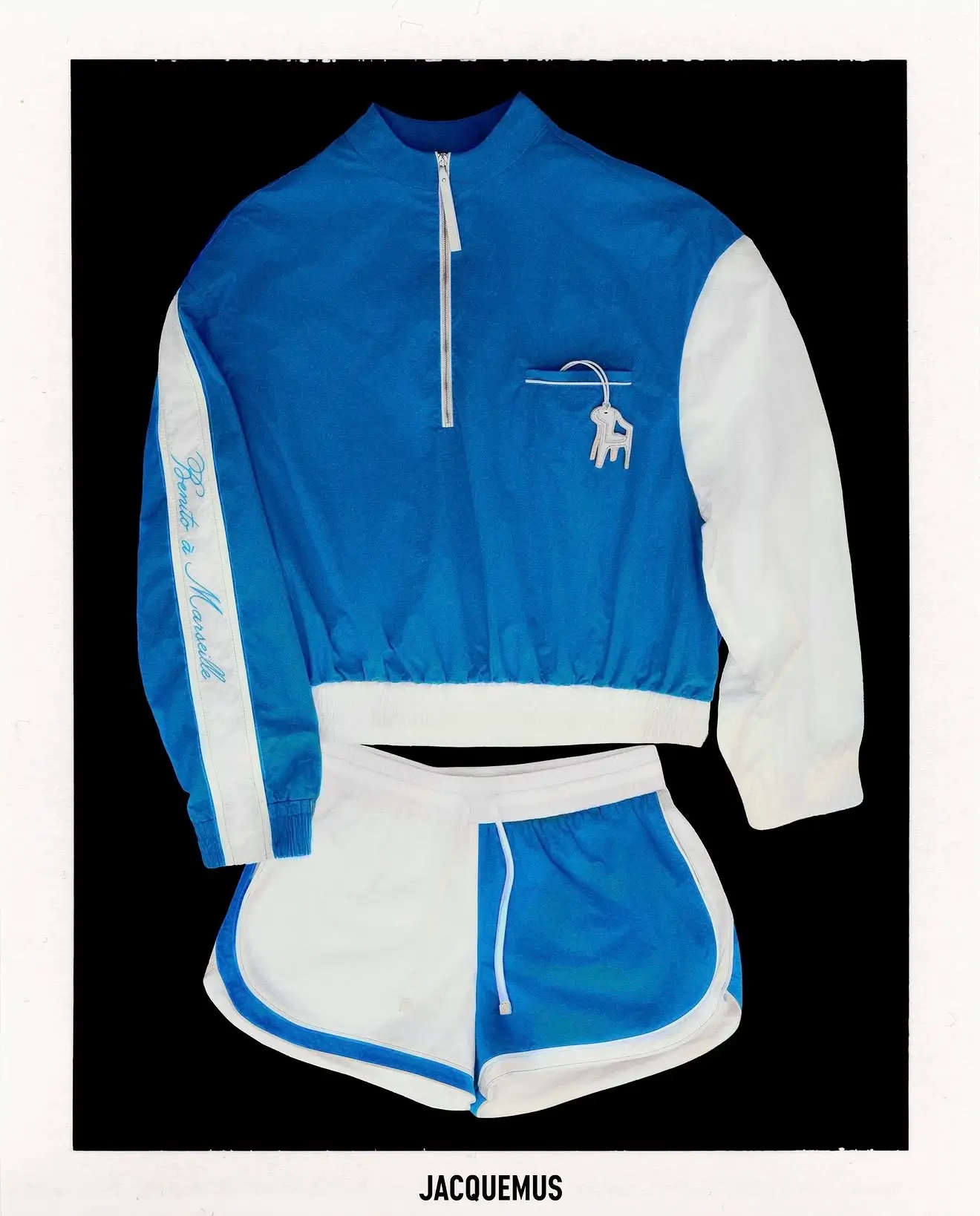 Front view of a blue-and-white Jacquemus athletic set featuring a quarter-zip windbreaker with contrasting sleeves, matching split-color running shorts, and signature branded details displayed against a black background.