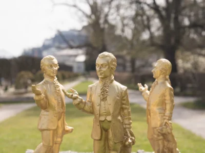 Three gold-painted Mozart statues stand on a dark stone pedestal in a park, each posed with one hand extended. One figure is accompanied by a small dog, while blurred trees and pathways create a soft-focus garden backdrop