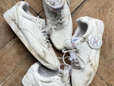 Four well-worn white Celine leather shoe are arranged in a circular pattern on a herringbone wood floor. The shoes feature visible scuffs, dirt marks, creases, and distressed soles, highlighting their heavily worn appearance. One sneaker displays a branded hangtag attached to the laces, while subtle Celine branding is visible on the tongue and insole. The overhead composition emphasizes the vintage, lived-in aesthetic and premium leather construction