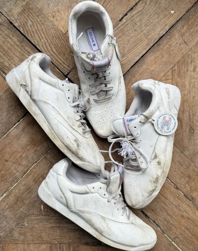 Four well-worn white Celine leather shoe are arranged in a circular pattern on a herringbone wood floor. The shoes feature visible scuffs, dirt marks, creases, and distressed soles, highlighting their heavily worn appearance. One sneaker displays a branded hangtag attached to the laces, while subtle Celine branding is visible on the tongue and insole. The overhead composition emphasizes the vintage, lived-in aesthetic and premium leather construction