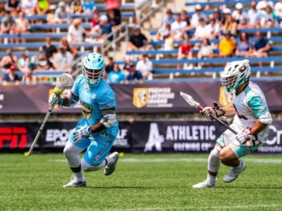 A lacrosse player in a light blue uniform sprints across the field while cradling the ball in his stick, closely pursued by a defender in a white and teal uniform. Both athletes wear helmets, gloves, and protective pads as they battle for possession during a professional outdoor game. A referee stands in the background, with spectators filling the stadium bleachers and sponsor banners lining the sidelines under bright daylight