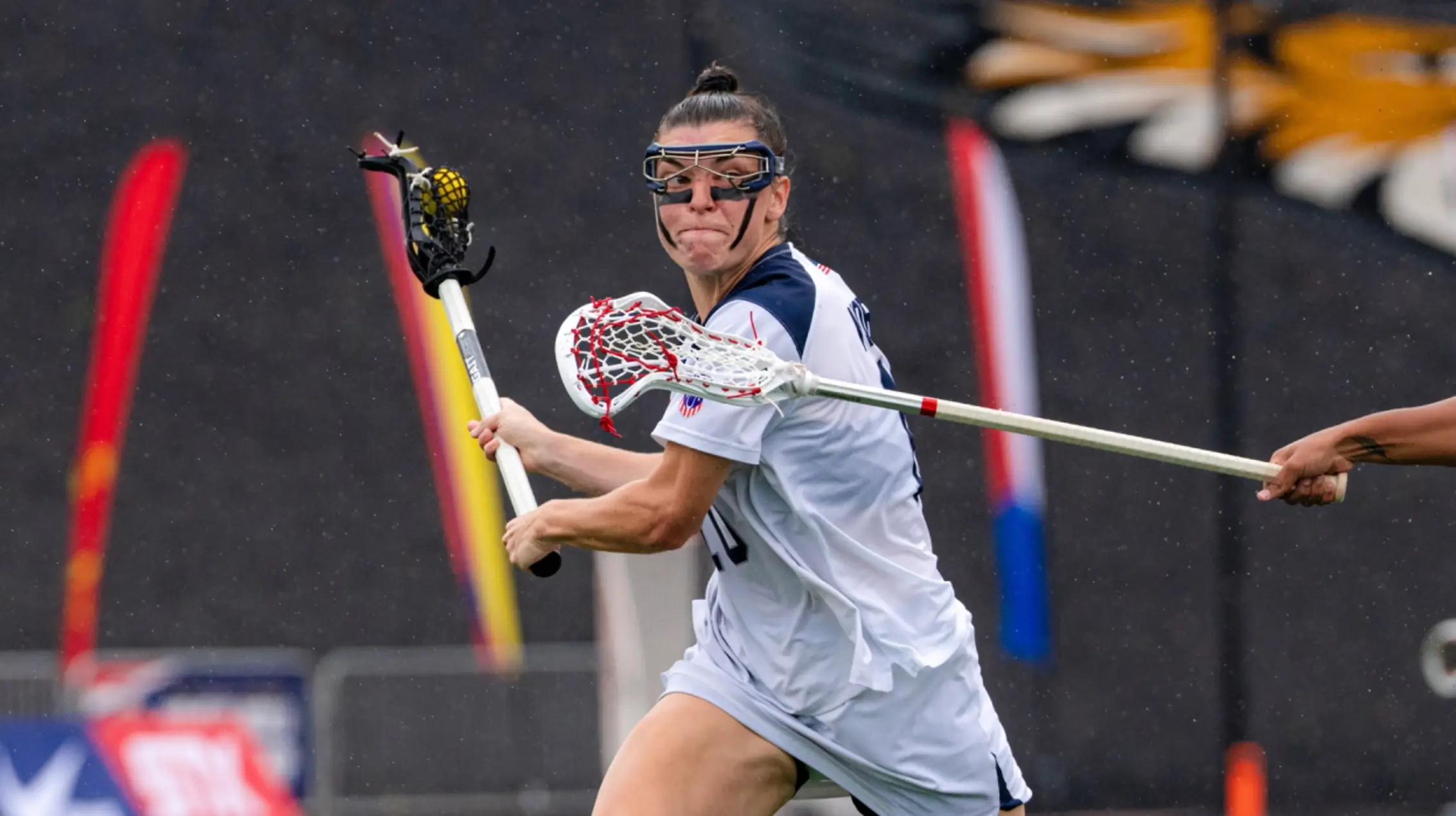 Women's lacrosse player in a white jersey and protective goggles cradles a yellow ball while preparing to pass as an opponent's stick challenges the play during an outdoor match.