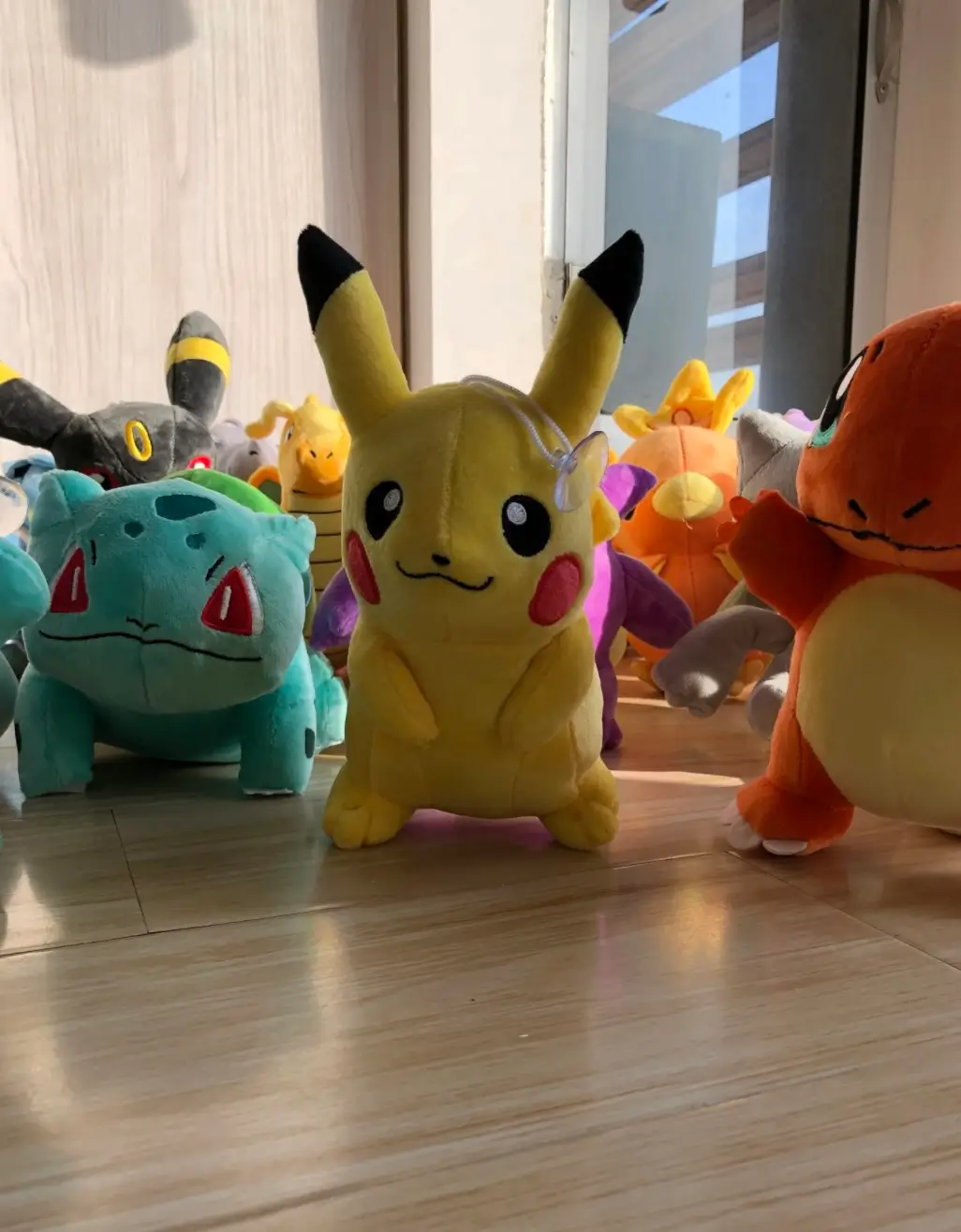Collection of Pokémon plush toys gathered on a sunlit wooden floor, with Pikachu standing at the center surrounded by Bulbasaur, Charmander, Umbreon, and other fan-favorite characters in a playful group display.