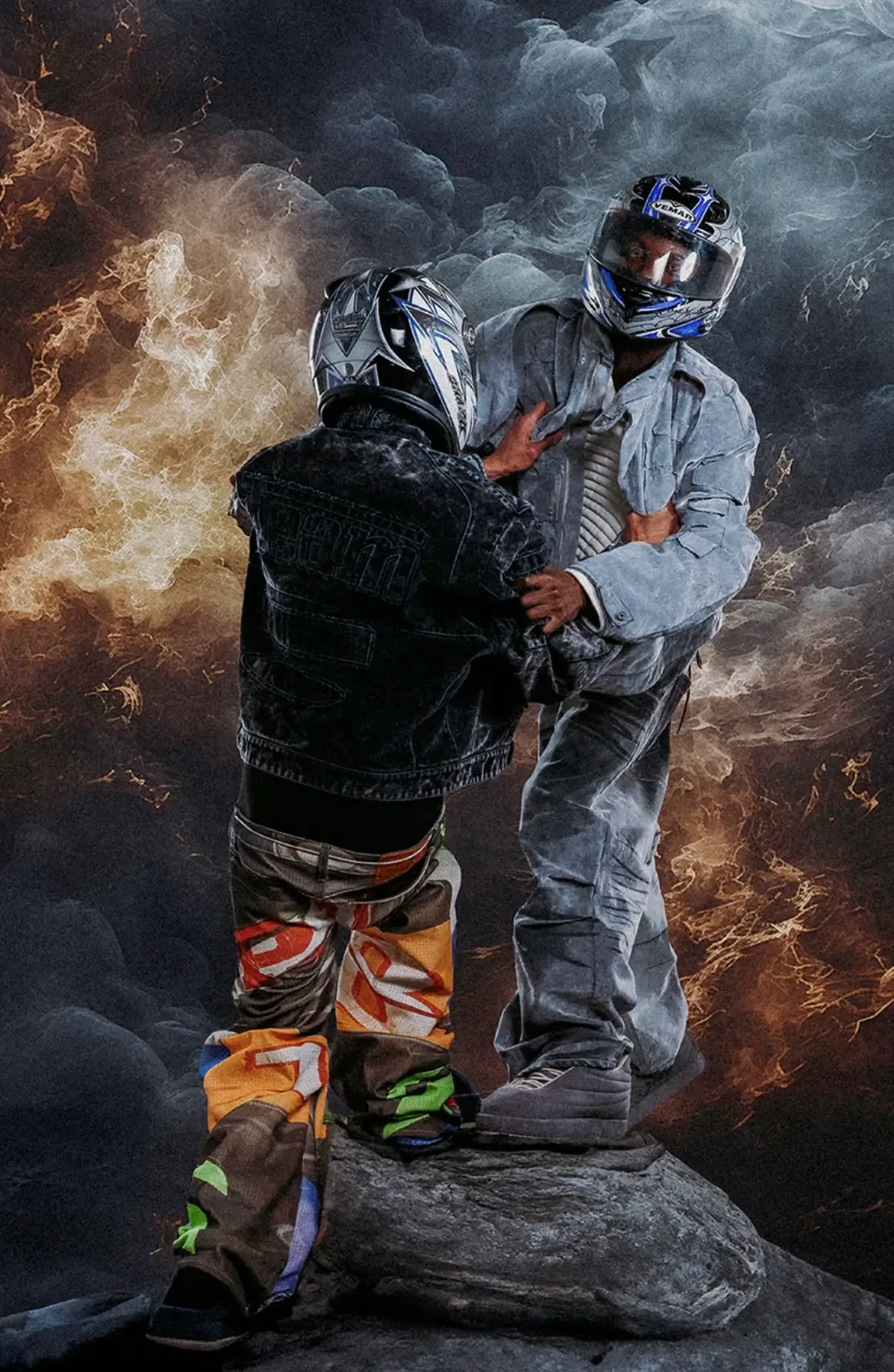 Two models wearing oversized denim outfits and full-face motorcycle helmets pose dramatically atop a rock, surrounded by swirling smoke and fiery visual effects, creating a cinematic campaign for a contemporary streetwear collection.