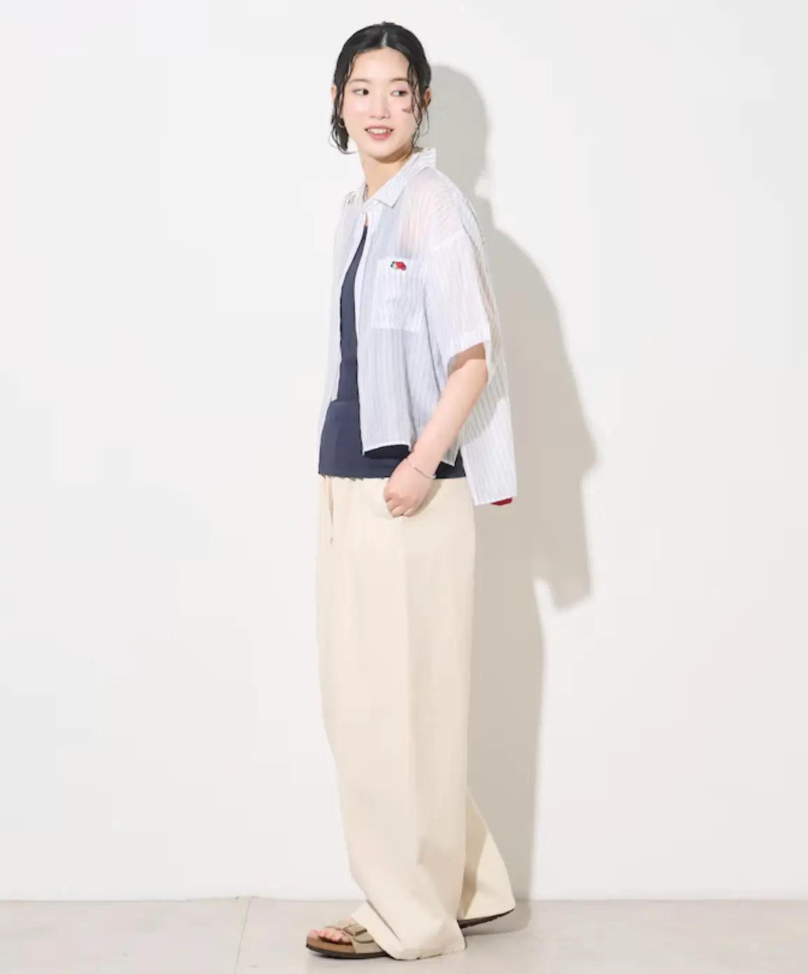 Model wearing a sheer white striped short-sleeve shirt from the FREAK'S STORE × Fruit of the Loom collaboration over a dark tank top, styled with wide cream trousers and sandals.