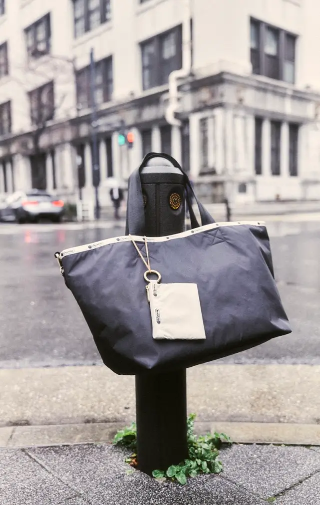 Large navy LeSportsac tote bag with cream trim and matching zip pouch displayed on a city bollard along a rain-soaked street, highlighting the collection's lightweight urban travel aesthetic.