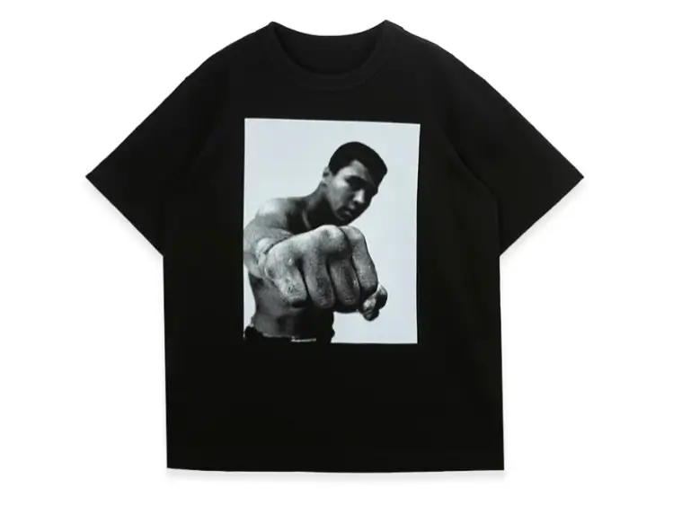 Black T-shirt featuring an archival black-and-white Muhammad Ali photograph with the boxing legend extending a punch toward the camera, presented as a bold front graphic on a minimalist crewneck silhouette.