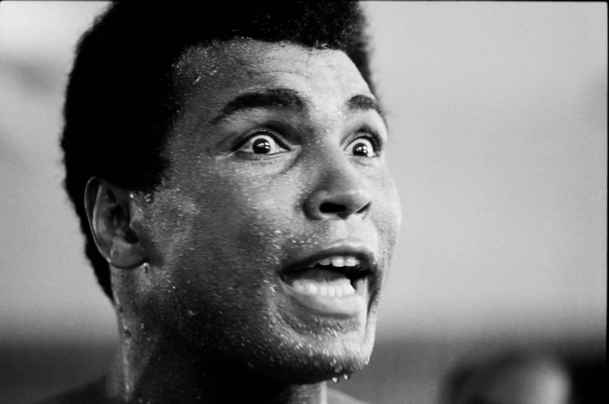 Black-and-white close-up portrait of Muhammad Ali during training, capturing his sweat-covered face, wide eyes, and animated expression in a candid moment that reflects his legendary charisma and intensity.