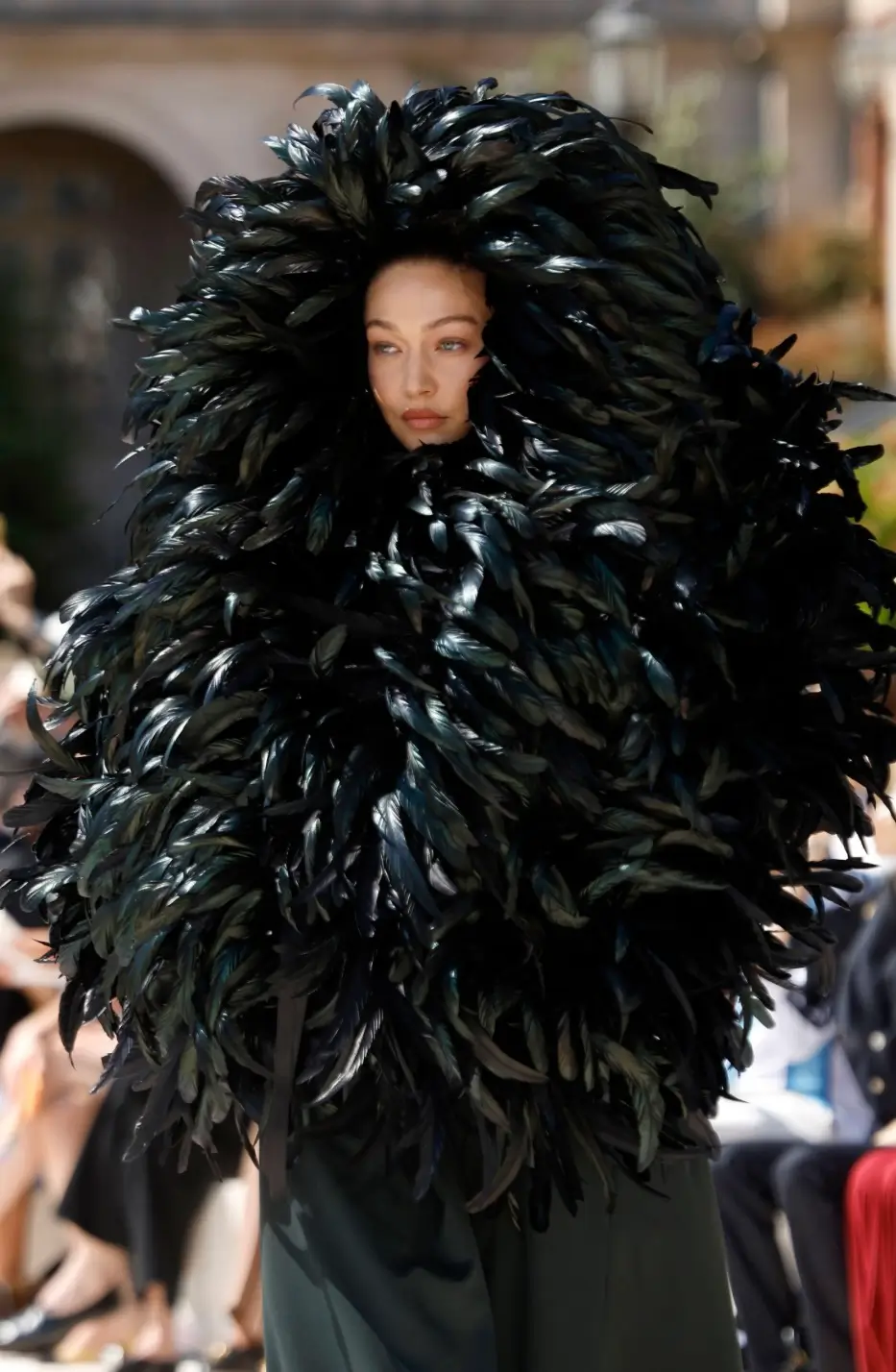 Runway model wearing an oversized black feathered couture ensemble with a sculptural hood framing the face during an outdoor fashion show.