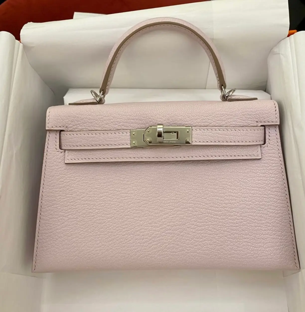 Pale pink Hermès Kelly handbag crafted from textured leather with palladium hardware, presented inside its original orange box with protective tissue paper.