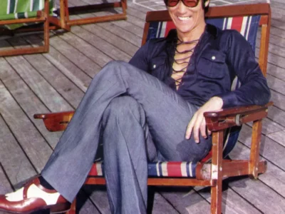 Vintage color photograph of Bruce Lee reclining in a striped wooden deck chair aboard a ship. He smiles broadly while wearing oversized amber-tinted aviator sunglasses, a deep navy lace-up shirt with an open neckline, tailored gray flared trousers, and two-tone brown-and-white leather shoes. Wooden deck planks, colorful folding chairs, lifebuoys, and ship railings surround him, creating a relaxed holiday atmosphere that contrasts with his intense on-screen persona