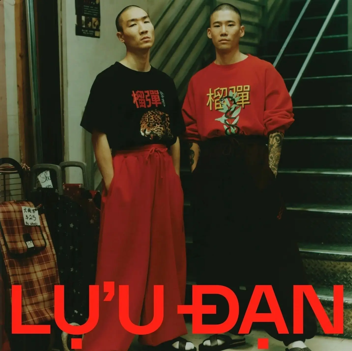 Two models stand in front of an indoor staircase wearing pieces from LỰU ĐẠN's collection. The model on the left wears a black graphic T-shirt featuring a leopard illustration and bold Chinese typography tucked into dramatic high-waisted red wide-leg trousers. The model on the right wears a red sweatshirt with large Chinese characters and a coiled snake graphic paired with oversized black trousers, revealing tattooed forearms. Both wear black slide sandals and shaved hairstyles, while the image is finished with a large red LỰU ĐẠN logo across the bottom, emphasizing the label's bold fusion of East Asian graphics, oversized tailoring, and underground streetwear aesthetics.