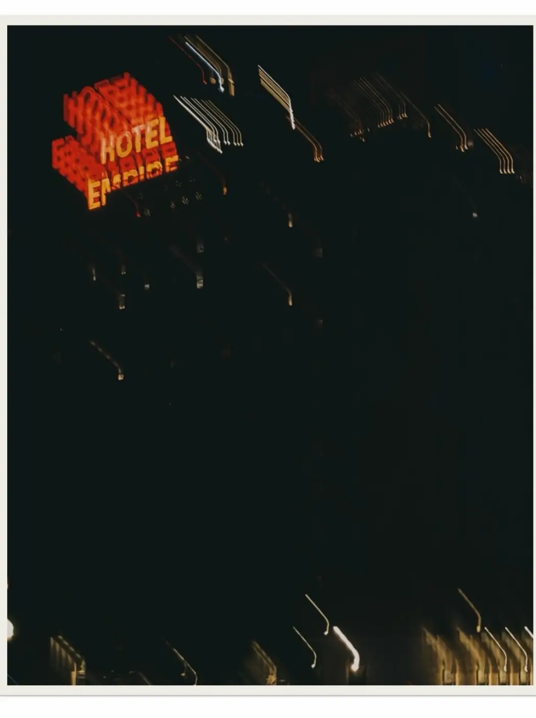 Blurred nighttime photograph of the Hotel Empire neon sign glowing in red and orange against a dark city skyline. Long-exposure camera movement creates streaks of light across the building, producing an abstract, dreamlike urban scene with minimal visible architectural detail.