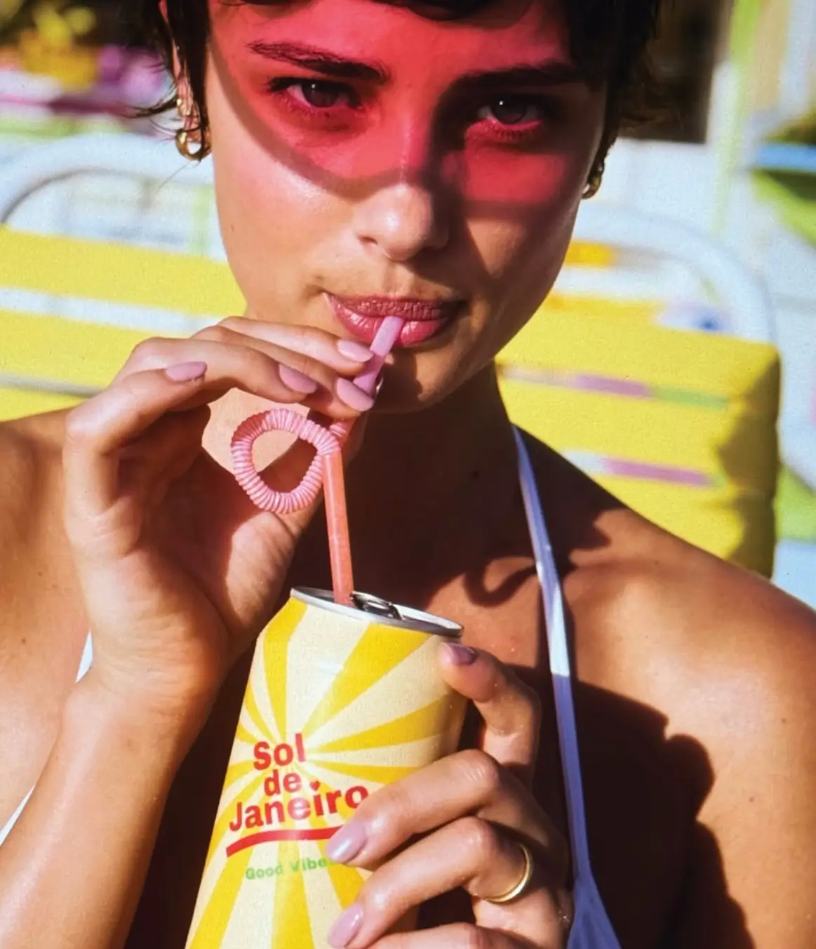 Close-up portrait of a woman in bright sunlight sipping from a pastel pink curly straw inserted into a yellow Sol de Janeiro branded drink can. Wearing a white halter swimsuit and gold jewelry, the subject is partially shaded by a vivid pink visor, with colorful lounge chairs softly blurred in the background for a playful, summer-inspired scene.