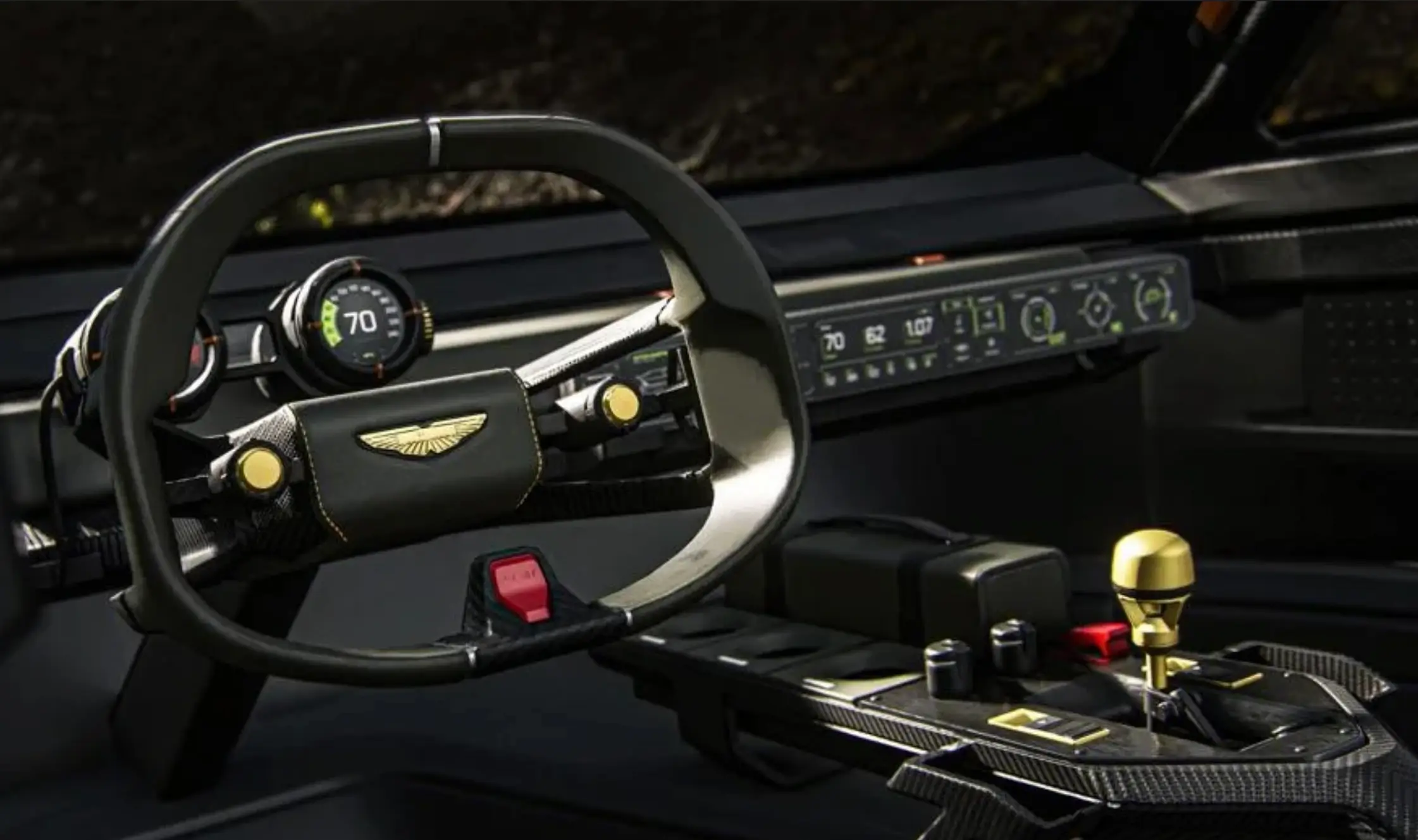 Close-up of the Aston Martin Dreadnought concept interior, featuring a minimalist steering wheel with the Aston Martin wing emblem, a circular digital driver's display, a full-width dashboard interface, exposed carbon-fiber trim, metallic gold switchgear, and a sculptural center console with a gold gear selector.