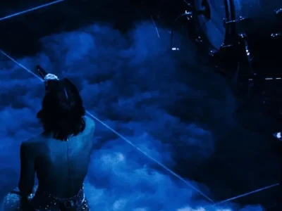 Grace Abrams sits on a dimly lit stage surrounded by low-lying fog, holding a microphone while facing away from the audience. Blue lighting washes over the scene, with a drum kit visible nearby, creating a moody, cinematic concert atmosphere