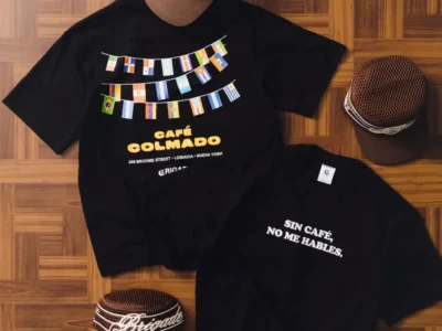 Flat-lay of a Café Colmado apparel collection arranged on a warm parquet wood floor. A black graphic T-shirt features strings of colorful international flags above bold yellow "CAFÉ COLMADO" lettering, with the address "266 Broome Street · Loisaida · Nueva York" printed below. Beside it is a second black T-shirt with the white Spanish phrase "SIN CAFÉ, NO ME HABLES." Three brown mesh trucker caps with white embroidered script branding are positioned around the shirts, creating a coordinated merchandise display with earthy tones and strong graphic contrast