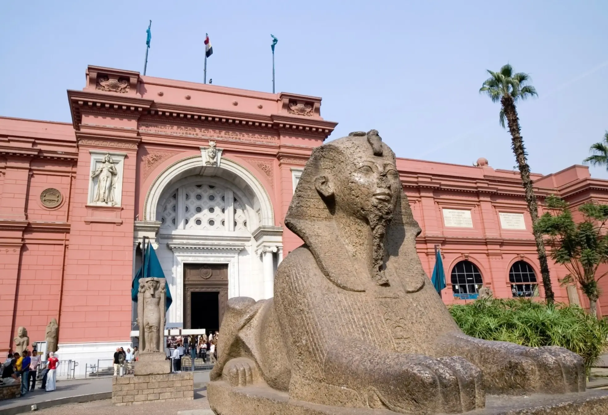 Ancient Egyptian sphinx statue positioned outside the Egyptian Museum in Cairo, with the museum's iconic pink neoclassical façade, palm trees, and visitors gathered near the main entrance.