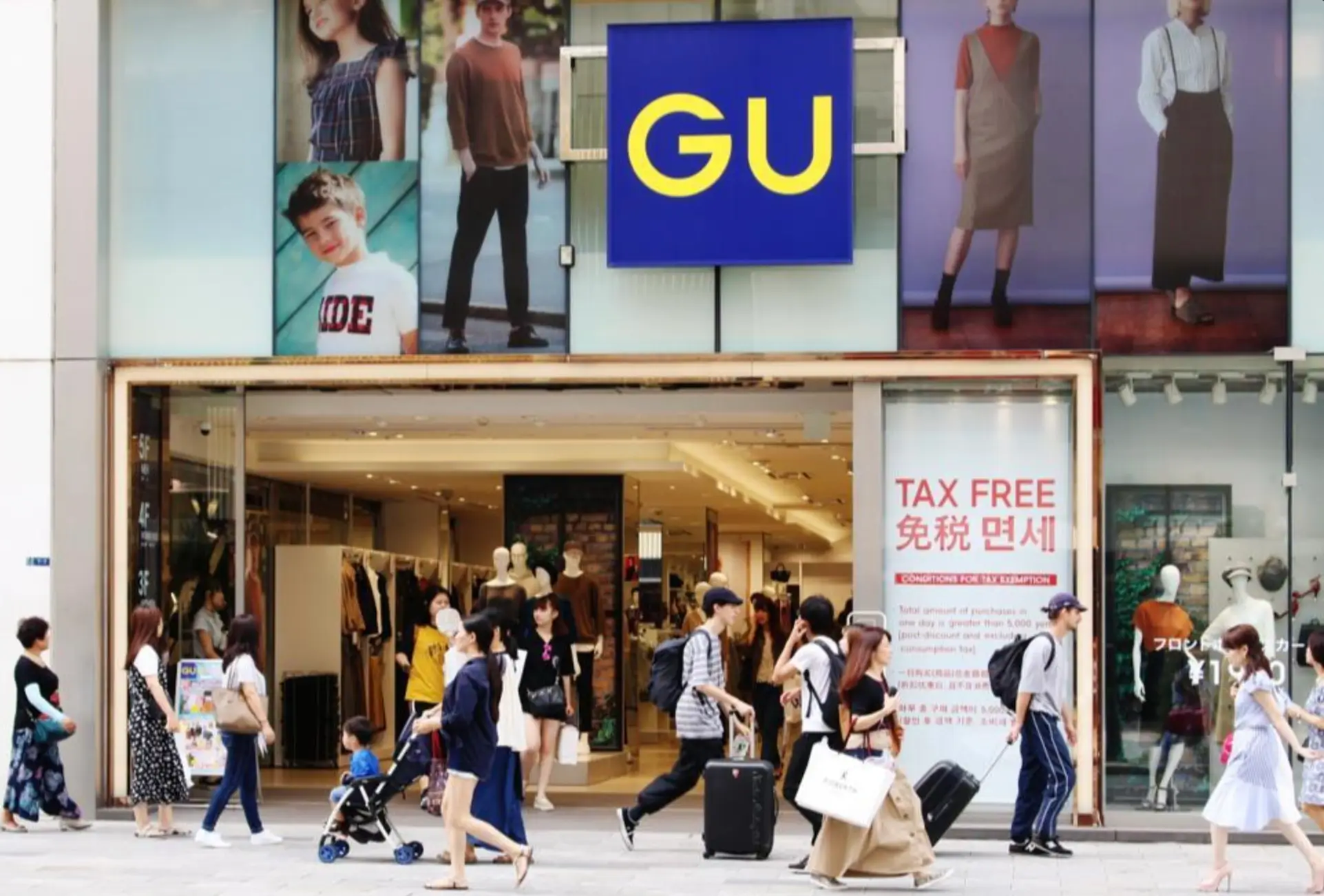 Street-level view of a GU retail store with the brand's blue-and-yellow logo above the entrance. Shoppers carrying bags and rolling suitcases walk past the storefront, while large fashion campaign images and a prominent tax-free shopping sign frame the busy urban scene.