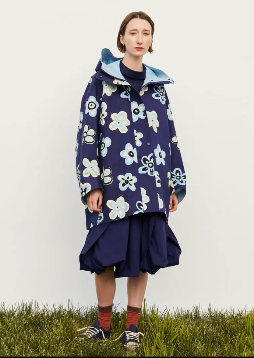 Model stands on grass against a plain off-white background wearing an oversized navy hooded coat covered in pale blue and cream floral graphics, layered over a sculptural navy skirt with striped socks and dark lace-up shoes.