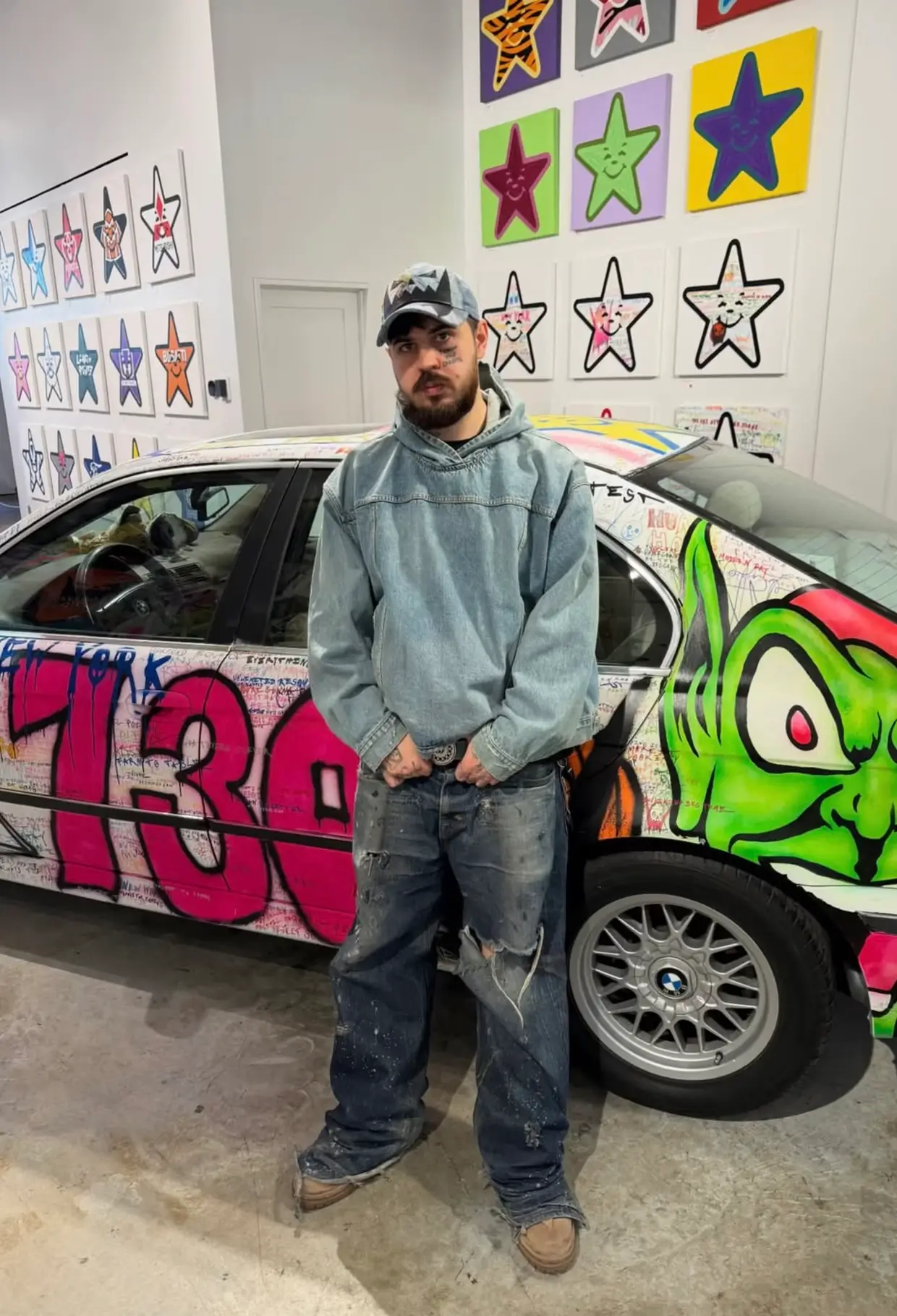 Artist standing inside a gallery beside a graffiti-covered BMW, surrounded by colorful star paintings displayed across the walls.