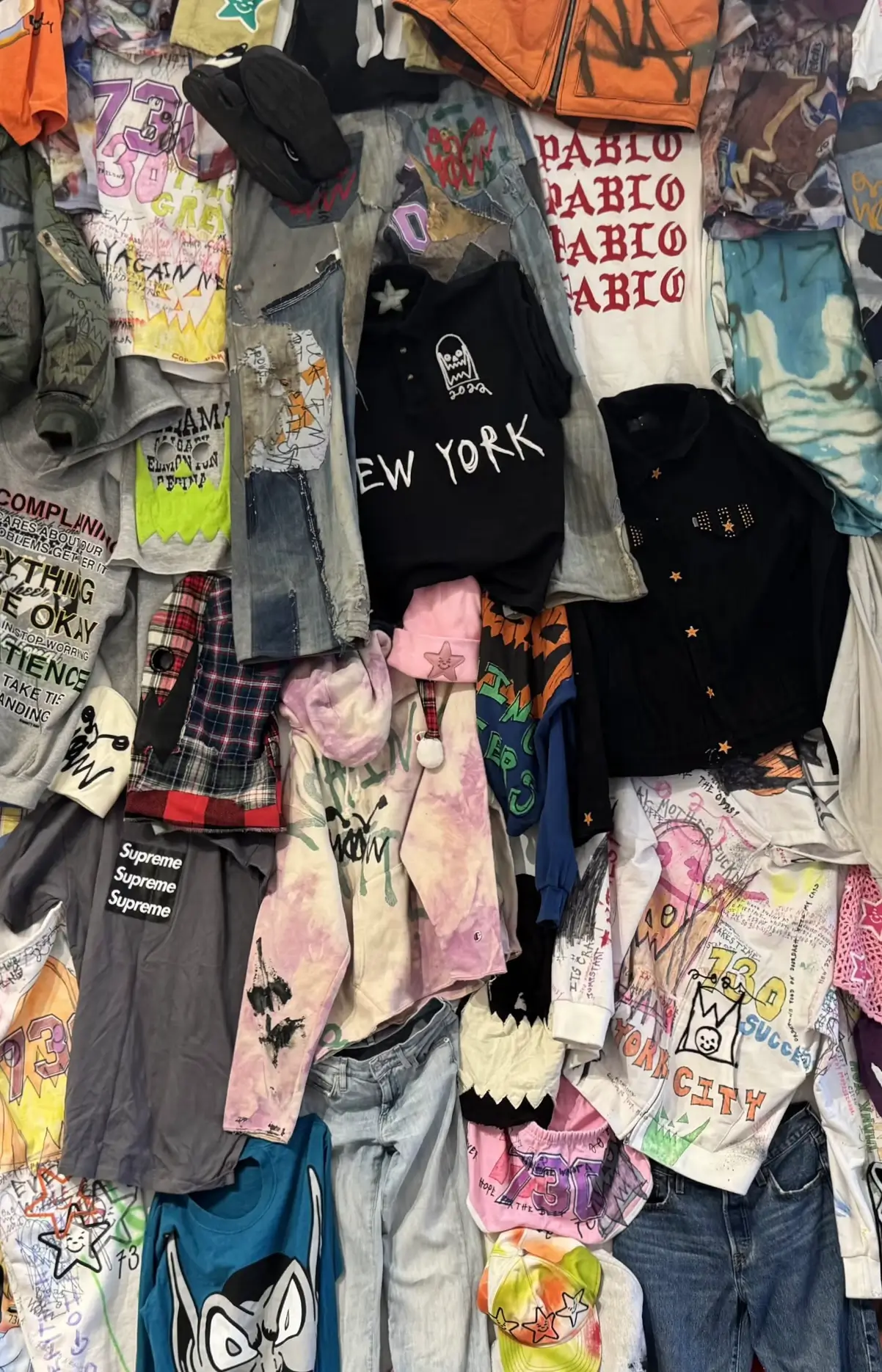Wall installation displaying dozens of custom streetwear garments, including hand-painted hoodies, graphic T-shirts, distressed denim, jackets, shorts, and accessories covered in graffiti, doodles, stars, and bold lettering.