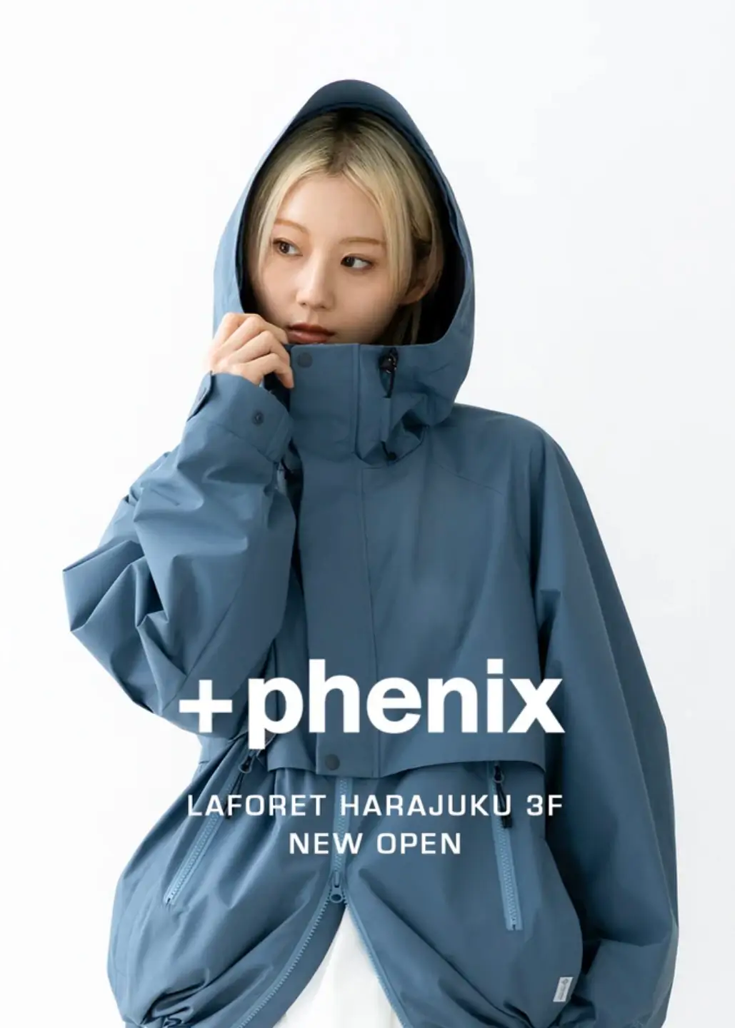 Model wearing a blue-gray +phenix technical hooded shell jacket with a high storm collar, waterproof zipper details, and a relaxed silhouette in a minimalist studio.