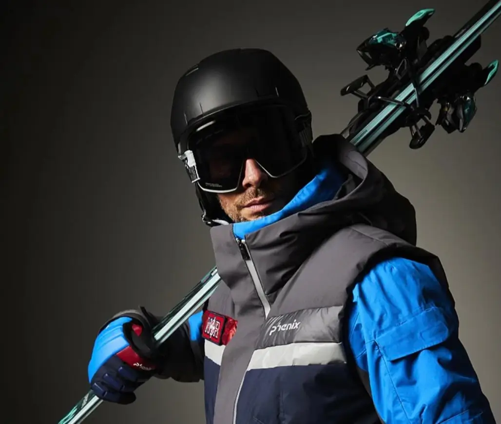 Skier wearing a black helmet, oversized goggles, blue technical jacket, and insulated Phenix vest while carrying skis across one shoulder.