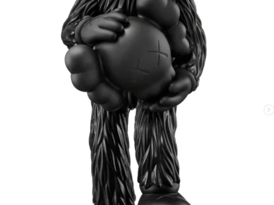 Black KAWS collectible figure standing headless while cradling its detached Companion head, with the severed neck revealing a bright red tree-ring cross section and a detached hand resting beneath one foot against a white background