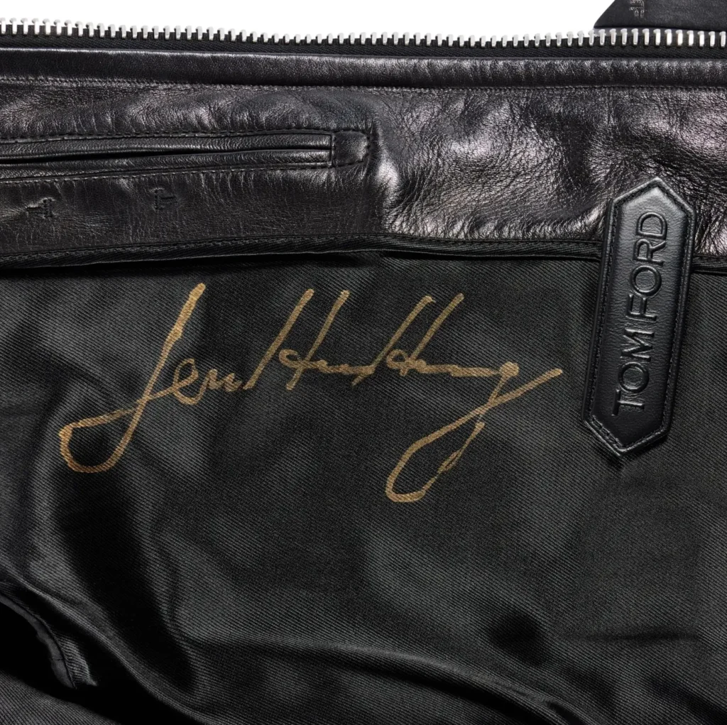 Close-up of the interior lining of a black Tom Ford leather jacket featuring Jensen Huang's gold signature across the black fabric. A black leather Tom Ford zipper pull sits beside the autograph