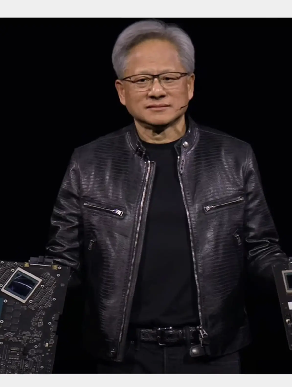 Jensen Huang stands onstage wearing a black crocodile-embossed leather jacket over a black T-shirt while presenting NVIDIA hardware. He holds two large graphics processing boards against a dark stage backdrop, highlighting the jacket's stand collar, silver zipper, and horizontal textured pattern.