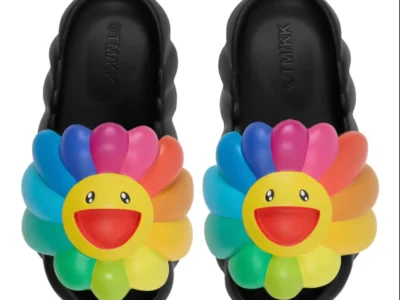 Pair of black molded slide sandals viewed from above, each featuring a large multicolored smiling flower across the strap. The thick sculpted sole contrasts with the bright rainbow petals and yellow flower face