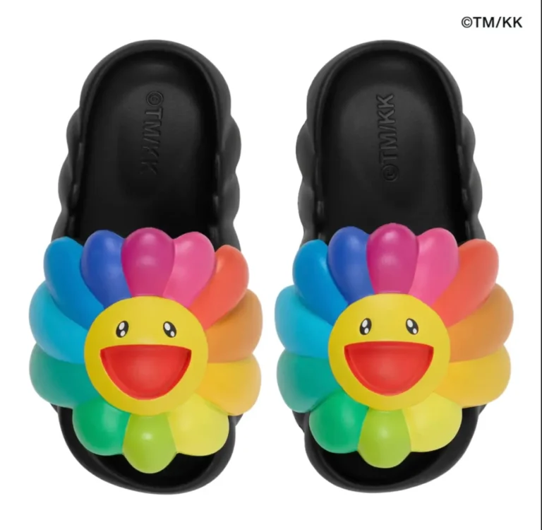 Pair of black molded slide sandals viewed from above, each featuring a large multicolored smiling flower across the strap. The thick sculpted sole contrasts with the bright rainbow petals and yellow flower face