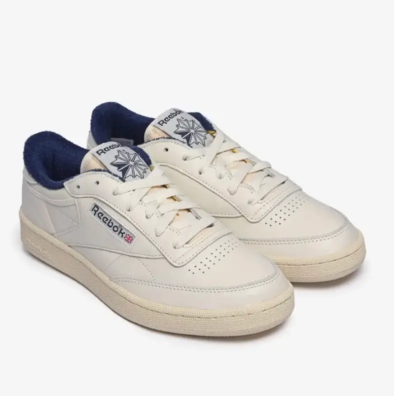 Pair of Reebok Club C low-top sneakers in off-white leather with vintage cream midsoles and deep navy terry cloth lining. The shoes feature classic Reebok side logos with Union Jack detailing, perforated toe boxes, white flat laces, and retro tongue labels, photographed against a clean light gray studio background.