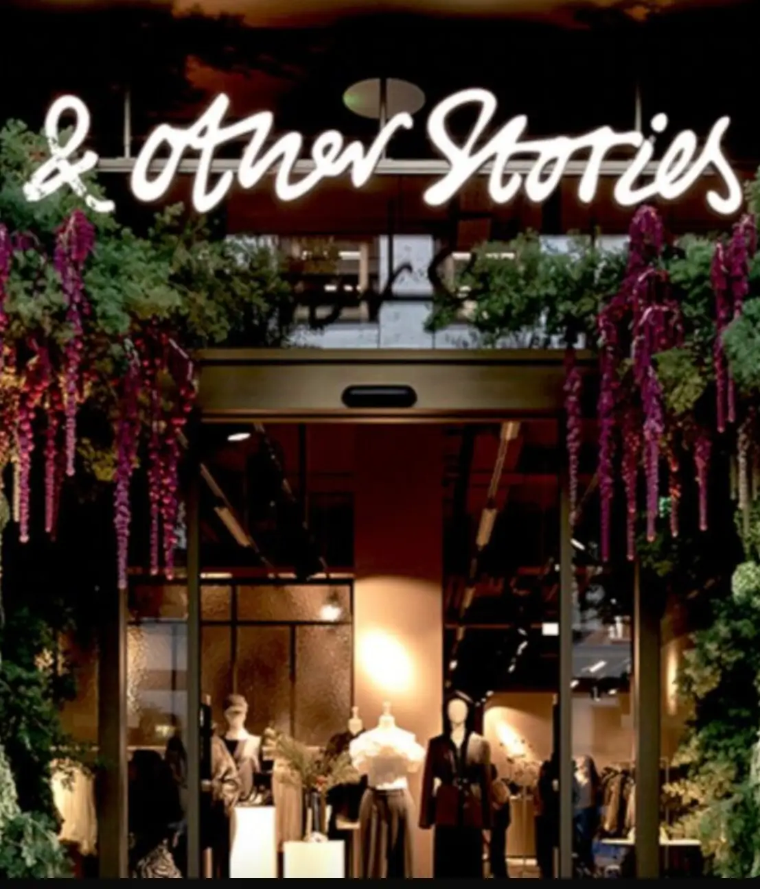 Exterior of an & Other Stories boutique at night, framed by lush hanging greenery and purple flowers. Warm interior lighting reveals mannequins and clothing displays through the glass entrance beneath the illuminated store sign.