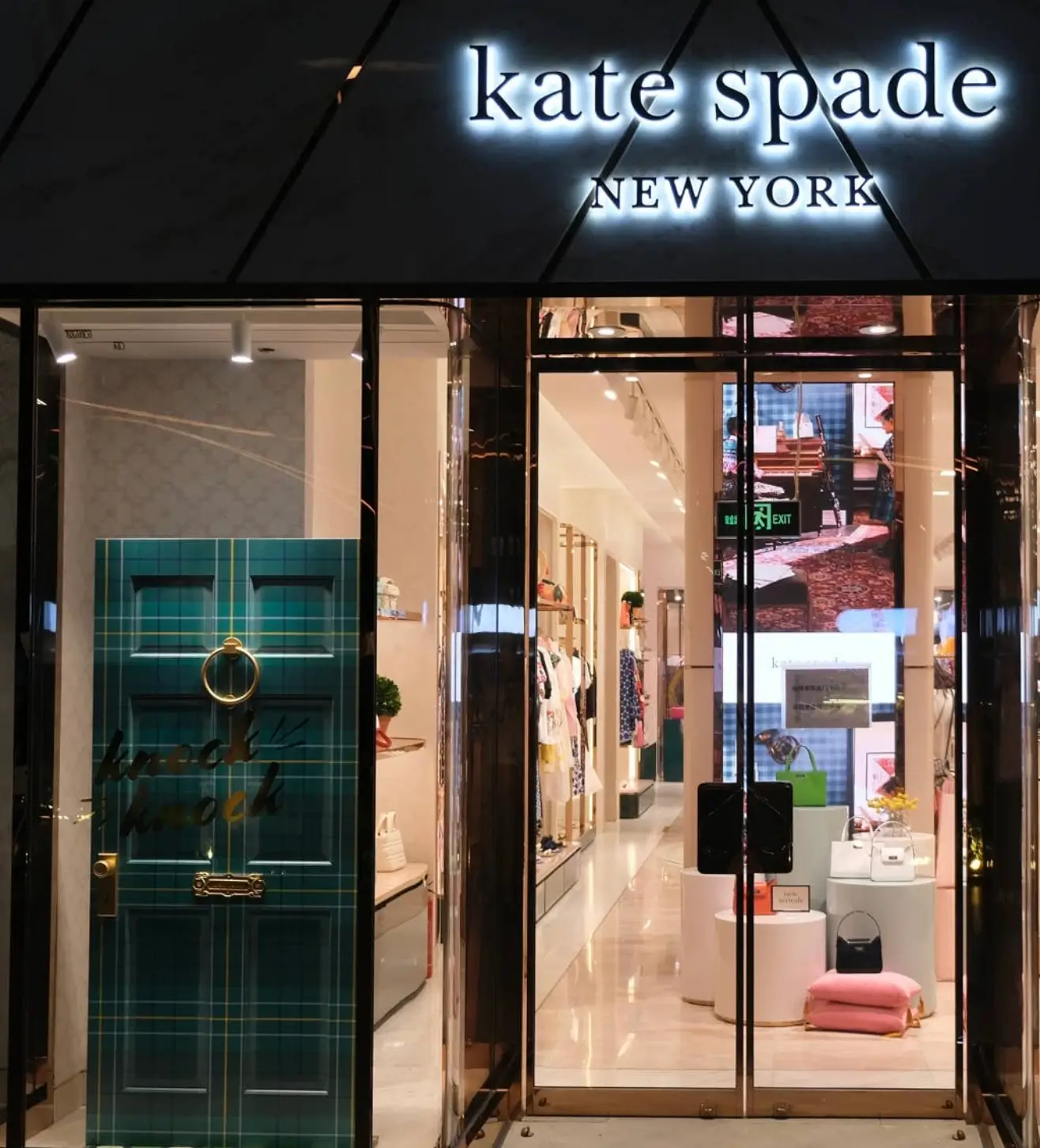Exterior of a Kate Spade New York boutique at night featuring an illuminated storefront sign, a distinctive green tartan display door, and a brightly lit interior showcasing handbags, accessories, and apparel.