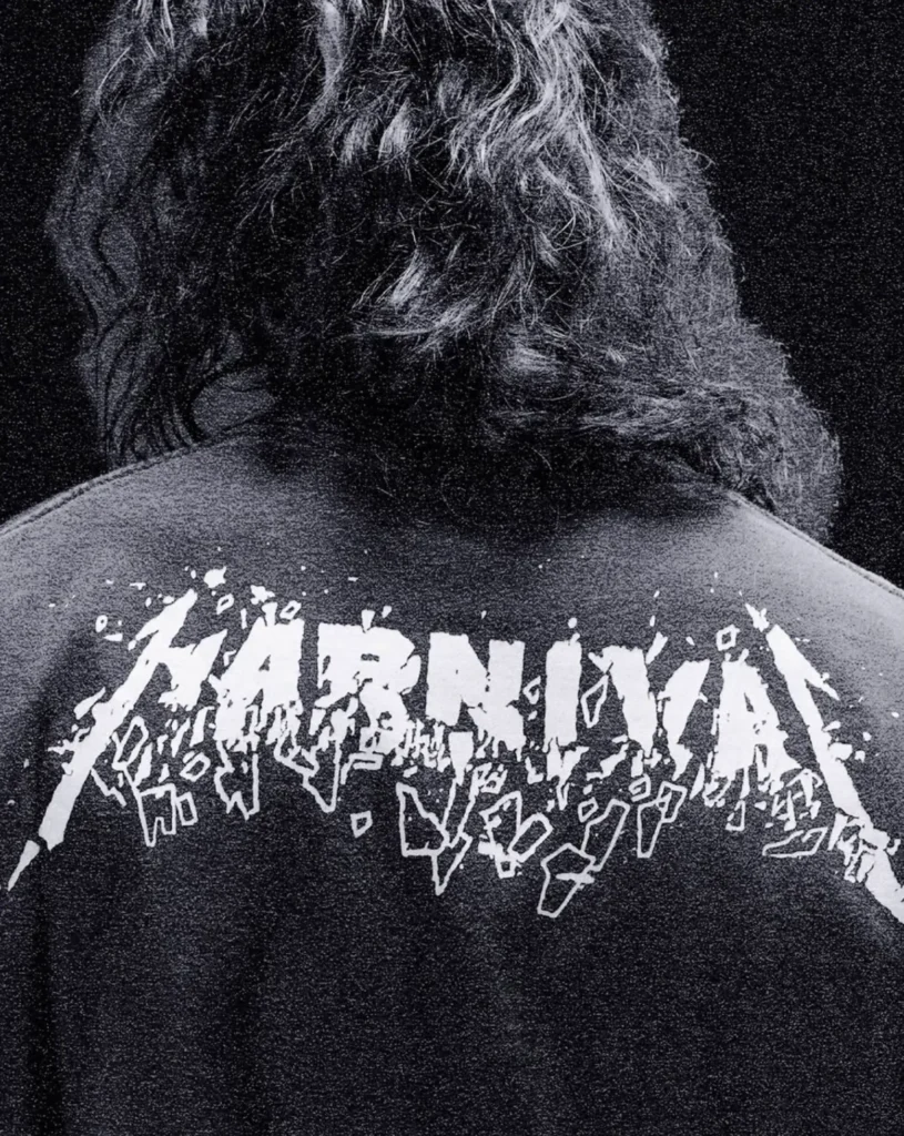 Back view of a person with shoulder length wavy hair wearing a black T-shirt featuring a distressed white Metallica logo across the upper back. The monochrome, grainy photograph emphasizes the bold graphic against a dark background