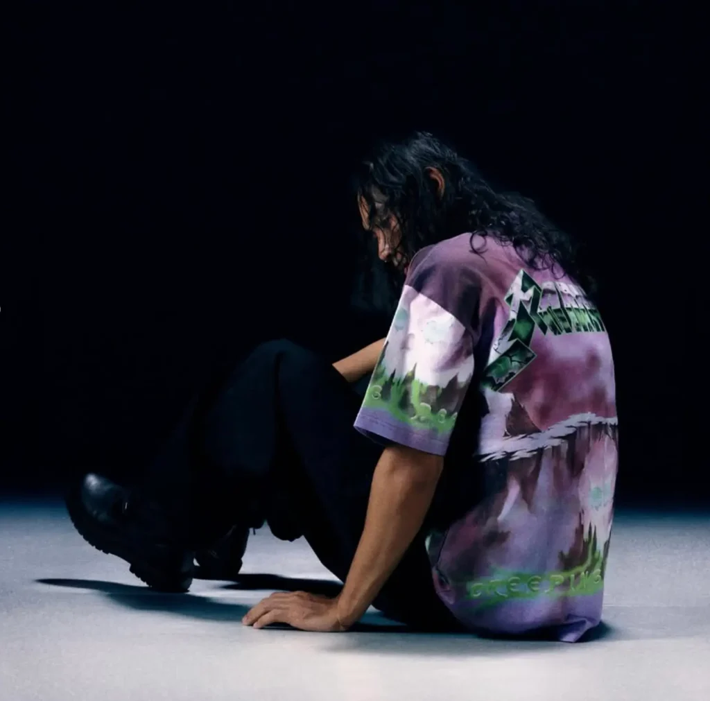 A model sits on the floor in a dark studio wearing a purple tie dye Metallica graphic T-shirt with a large green logo across the back and scenic artwork wrapping around the fabric. Loose black pants and black boots complete the relaxed style