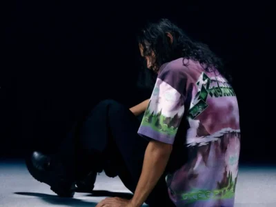 A model sits on the floor in a dark studio wearing a purple tie dye Metallica graphic T-shirt with a large green logo across the back and scenic artwork wrapping around the fabric. Loose black pants and black boots complete the relaxed style