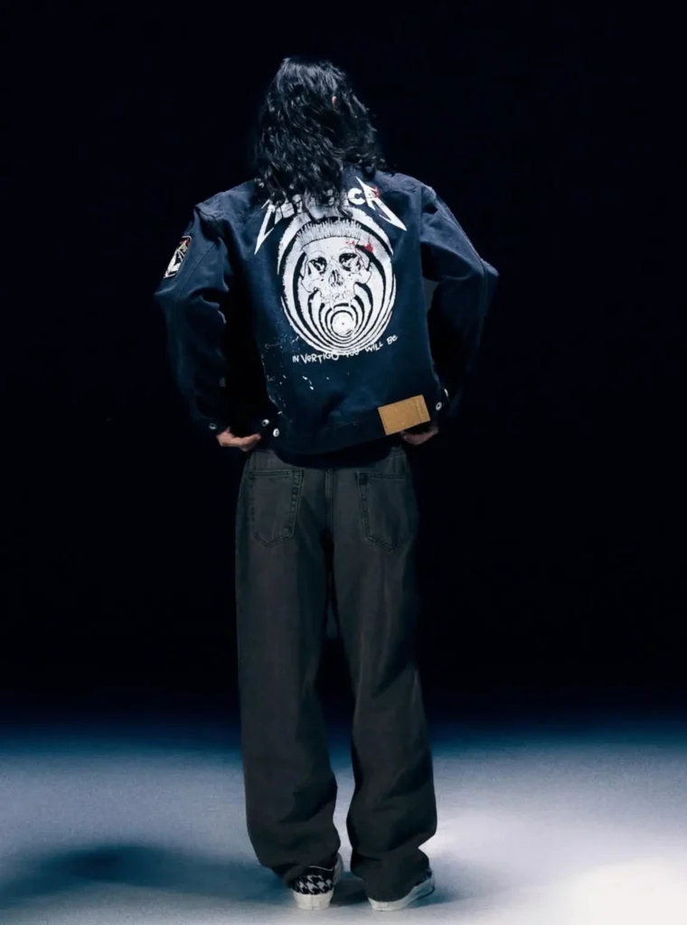 Rear view of a model wearing a dark denim jacket decorated with oversized Metallica artwork. A large circular skull graphic dominates the back panel beneath the band's logo, while the jacket is paired with loose charcoal jeans in a minimal studio set