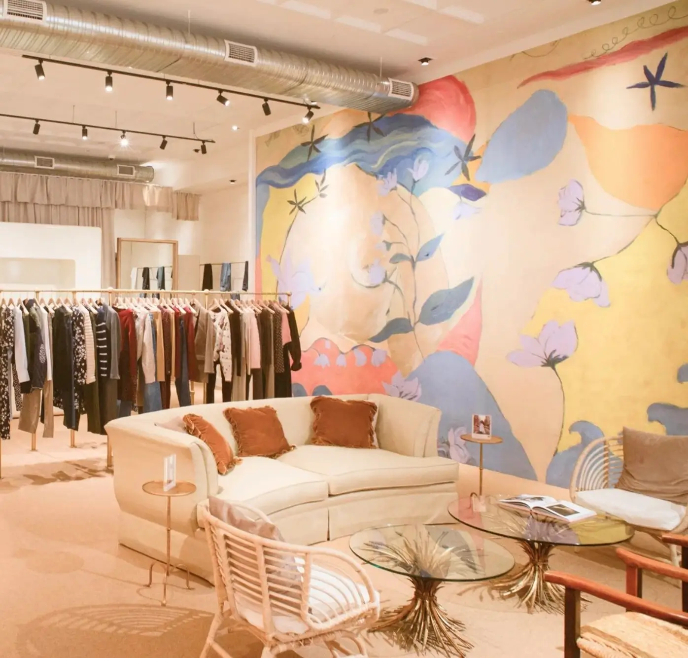 A boutique interior featuring a colorful hand-painted mural, cream lounge seating, glass coffee tables, and curated clothing racks under warm gallery-style lighting.