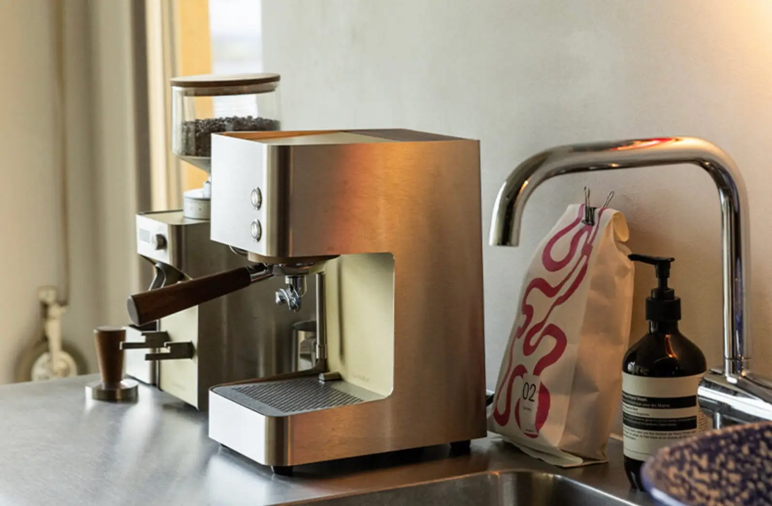 The ZURIGA E2-S espresso machine in a home kitchen beside a coffee grinder, coffee bag, and sink, showcasing its compact brushed stainless steel design in an everyday brewing setup.