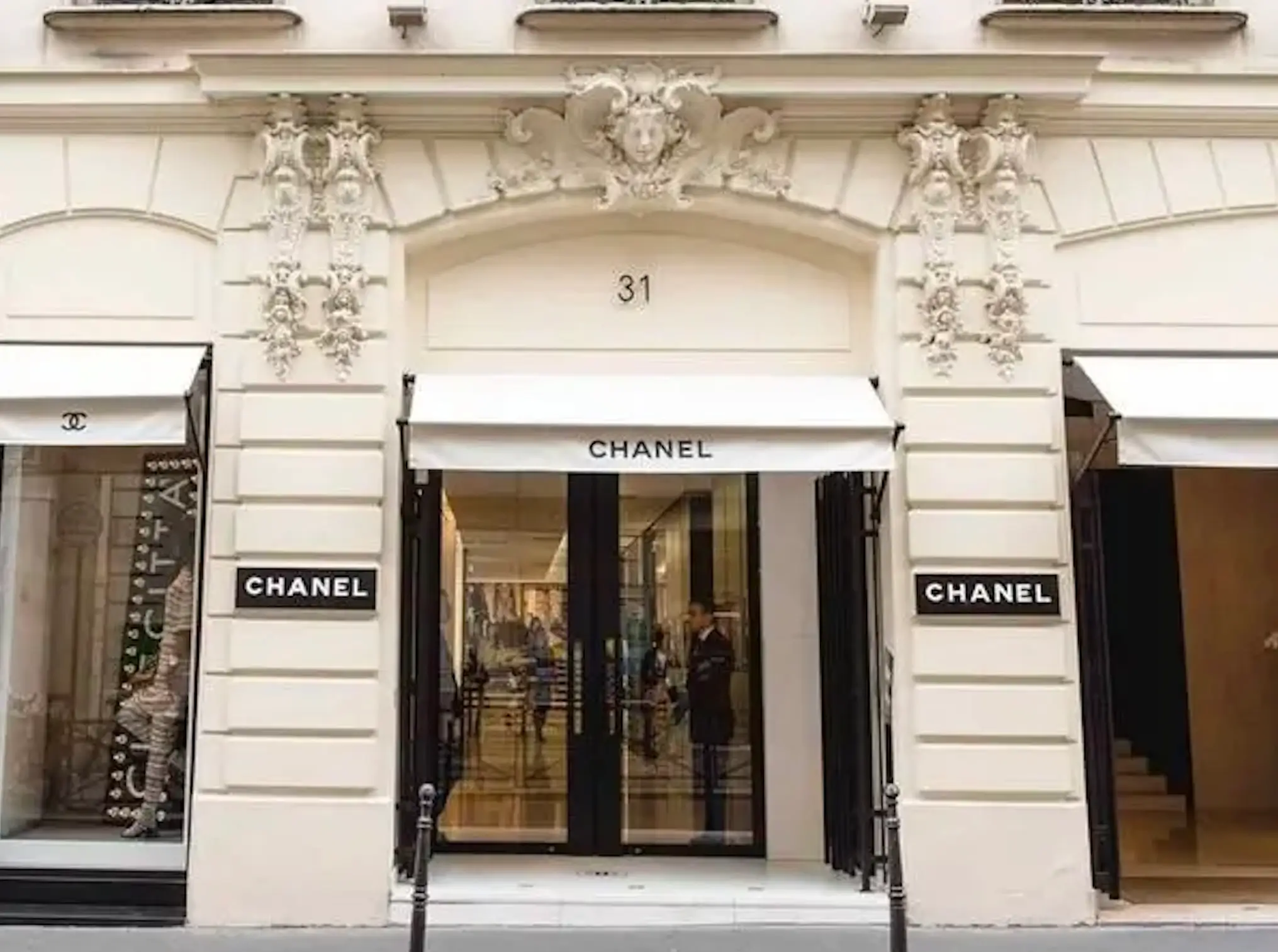 The exterior of Chanel's historic flagship boutique at 31 Rue Cambon in Paris, featuring the house's signature black and white storefront beneath an ornate Haussmann-style façade. Large display windows flank the central entrance, while cream stonework, decorative sculptural details, wrought-iron balconies, and classic Parisian architecture frame one of fashion's most iconic retail destinations.