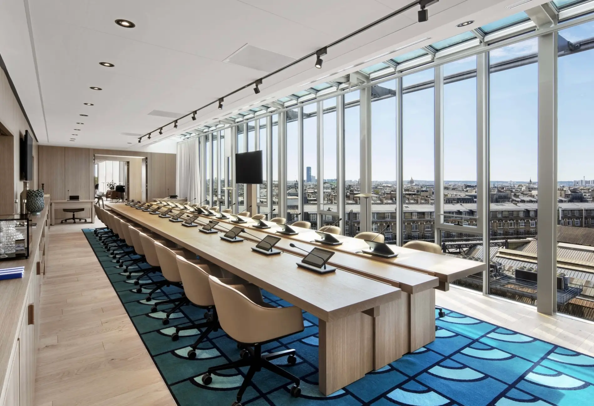 A contemporary executive boardroom inside Chanel's Paris headquarters, featuring a long light wood conference table equipped with touchscreens and microphones, surrounded by ergonomic chairs. Floor to ceiling glass windows flood the space with natural light while offering sweeping views across the Paris skyline, blending minimalist architecture with refined luxury.