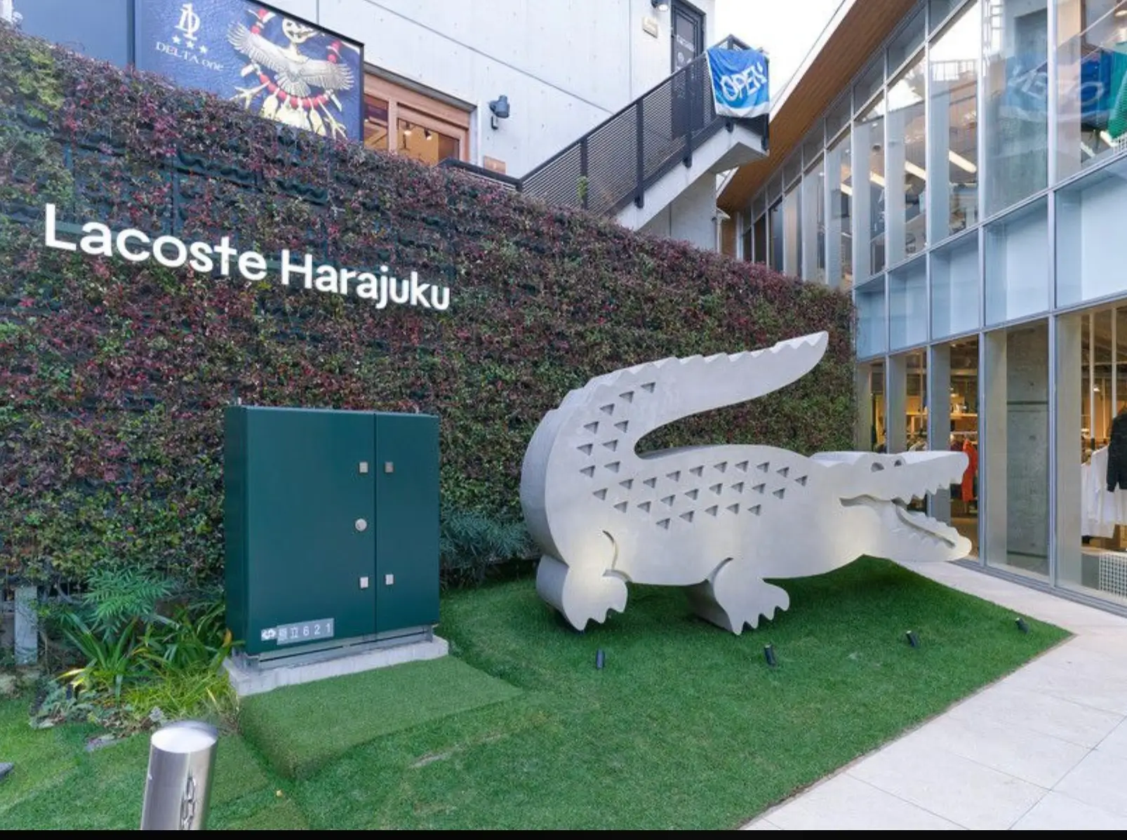 Exterior of Lacoste Harajuku in Tokyo featuring a large white crocodile sculpture displayed on a landscaped lawn beside the glass-fronted boutique. A green living wall with the Lacoste Harajuku sign forms the backdrop, while modern retail buildings and elevated walkways frame the entrance.