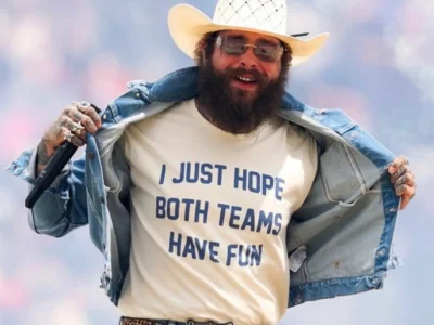 Post Malone wearing a white cowboy hat, tinted sunglasses, a light blue denim jacket, and blue jeans smiles on stage while holding a microphone and pulling open the jacket to reveal a cream T-shirt reading, “I JUST HOPE BOTH TEAMS HAVE FUN.” A leopard-print belt with a large silver buckle completes the outfit, with a softly blurred pastel concert backdrop behind him