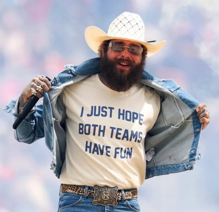 Post Malone wearing a white cowboy hat, tinted sunglasses, a light blue denim jacket, and blue jeans smiles on stage while holding a microphone and pulling open the jacket to reveal a cream T-shirt reading, “I JUST HOPE BOTH TEAMS HAVE FUN.” A leopard-print belt with a large silver buckle completes the outfit, with a softly blurred pastel concert backdrop behind him