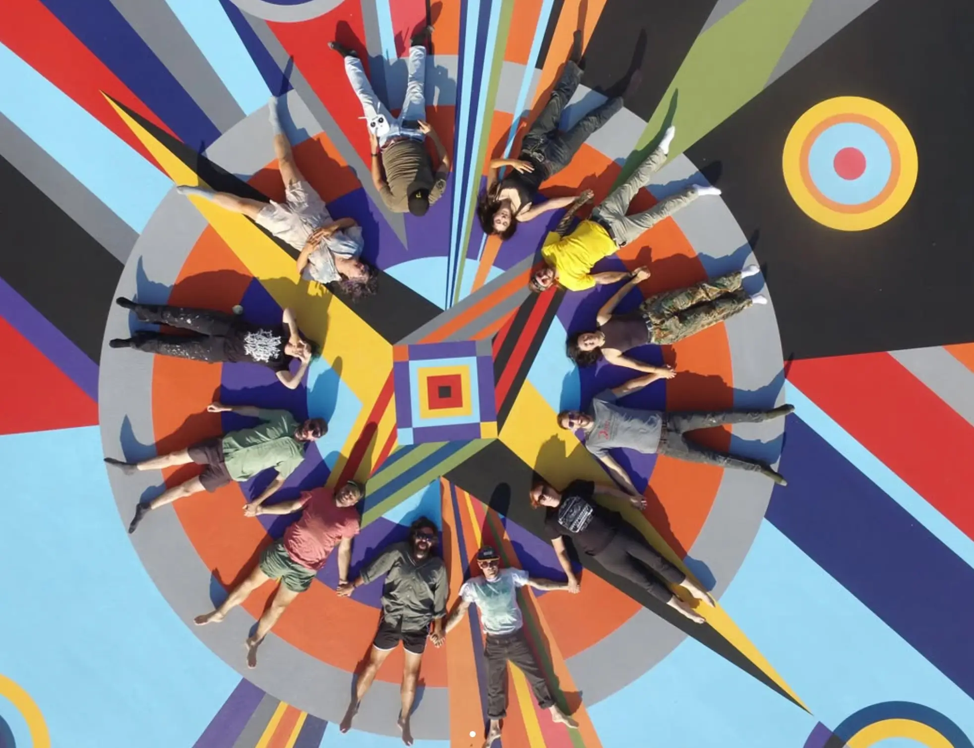 Overhead view of a group of people lying in a circle at the center of a vibrant geometric mural, holding hands around a colorful radial design painted across an outdoor sports court with bold stripes, circles, and contrasting shapes.